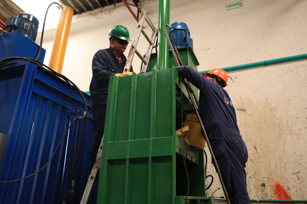 Dos hombres vestidos de azul, con casco naranja y verde, se encuentran sobre una escalera, manipulando una máquina.
