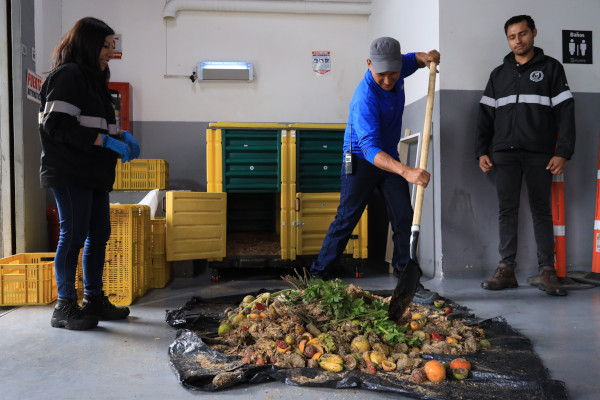 Un señor con una pala en su mano está mezclando residuos de frutas y verduras, mientras otro hombre con uniforme oscuro lo mira. En el fondo se ve una compostera.