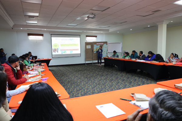 Grupo de personas reunidas en un auditorio. Las mesas están ubicadas en L, su mantel es de color naranja y las personas están observando la pantalla que proyecta la dinámica a desarrollar en la actividad. 