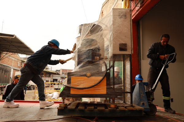 Es una fotografía de día. Y se puede ver dos hombres en plano general están sobre una grúa en la parte externa de una bodega, moviendo  maquinaria de procesamiento de plástico con un cargador de color azul.