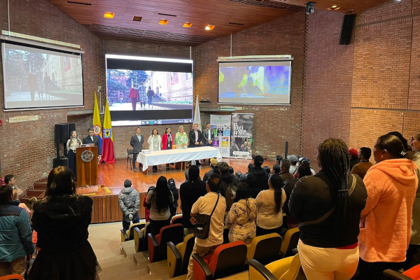 Foto al interior de un auditorio en contrapicada. Personas de pie en la mesa principal, el atril y sillas del auditorio. 