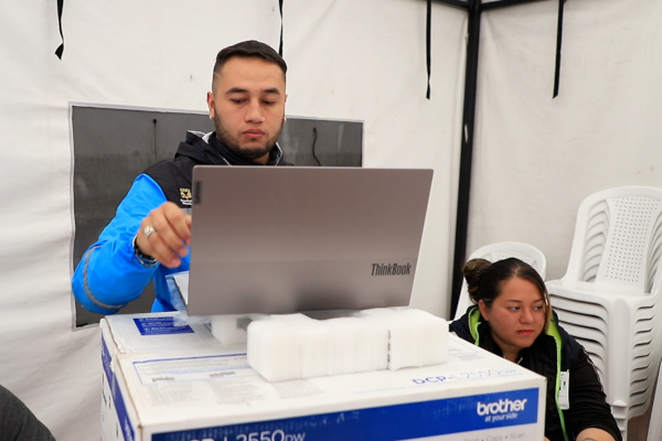 Foto al interior de una carpa blanca. En ella se ve a un joven de no más de 40 años, tiene puesta una chaqueta color azul cyan con negro y en sus manos tiene un computador portátil apoyado sobre una caja de impresora. Al lado derecho se ve a una mujer recicladora que está sentada y detrás de ella unas sillas de plastico, blancas.