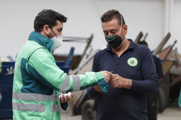 Foto en interior de un CTCC, en ella aparece un profesional de la UAESP con su chaqueta de dos ganas de verde, un reciclador/carretero que viste una camisa azul oscura está a su lado, recibiendo un carnet. En el fondo se ven carretas con material y un carretero se ve a espaldas.