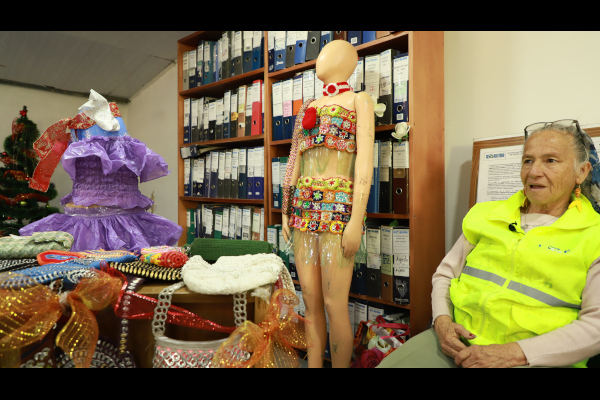  Foto en interiores. En la parte central derecha está una mujer, adulta mayor con un chaleco verde. Seguidamente están dos maniquíes con vestidos en material reciclable, en el fondo se ve un archivador con AZ.