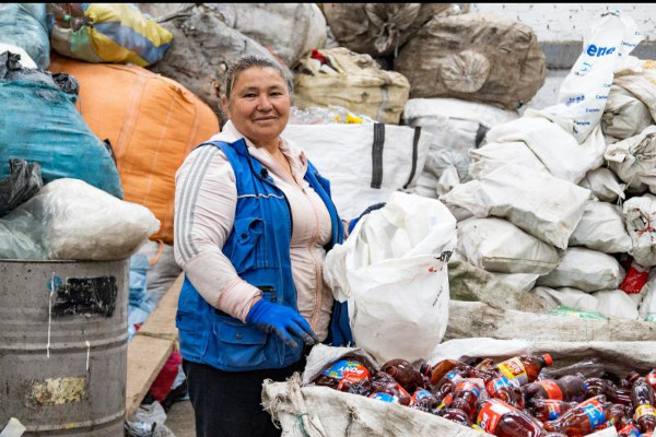 Foto de día en el interior de una bodega de reciclaje en la que se ven residuos almacenados en bolsas. En un plano 3/4 se ve a una mujer (representante legal de la Organización)  que está vestida con chaleco azul y sueter rosado y pantalón negro.