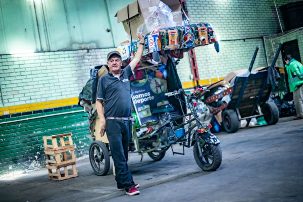 Foto en el interior de un CTCC Los Mártires. Reciclador vestido de color azul oscuro con una mano levantada sujetando su bicicarreta. En el fondo se ven residuos arrumados al fondo. Y una pared verde oscuro con verde claro