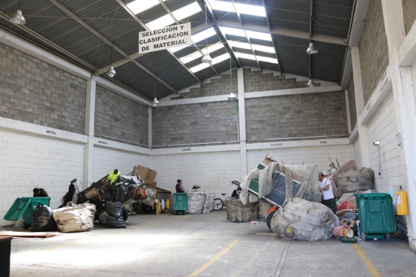 Foto de día, del interior de centro transitorio de Usaquén. Es una foto de día. Las paredes son con ladrllo y color blanco. El techo contempla tejas traslucidas. En el interior se encuentran 4 carreteros junto a sus carretas haciendo separación de material.