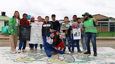 Niños y niñas encima del tapete didáctico posando para la foto con la bolsa negra y la bolsa blanca y el afiche del reciclador y la recicladora de oficio.