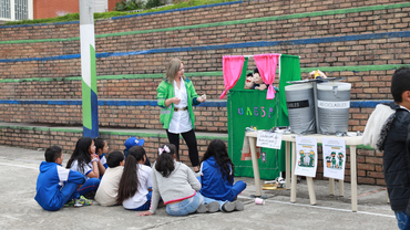 Stand de RBL con titiritero y dummies de la canica negra (NO RECICLABLES) y la canica blanca (RECICLABLES) los niños  y niñas mirando la presentación de títeres sentados en el suelo.