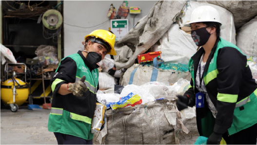 En la imagen: Dos recicladores en una bodega manipulan material reciclable. Uno de ellos, el del lado izquierdo, saluda cordialmente mostrando su pulgar hacia arriba.