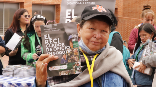 En la imagen: Una recicladora sostiene en sus manos un ejemplar del libro “El Reciclador de Oficio”. En el fondo, varias mujeres hacen fila  para recoger un ejemplar del libro.