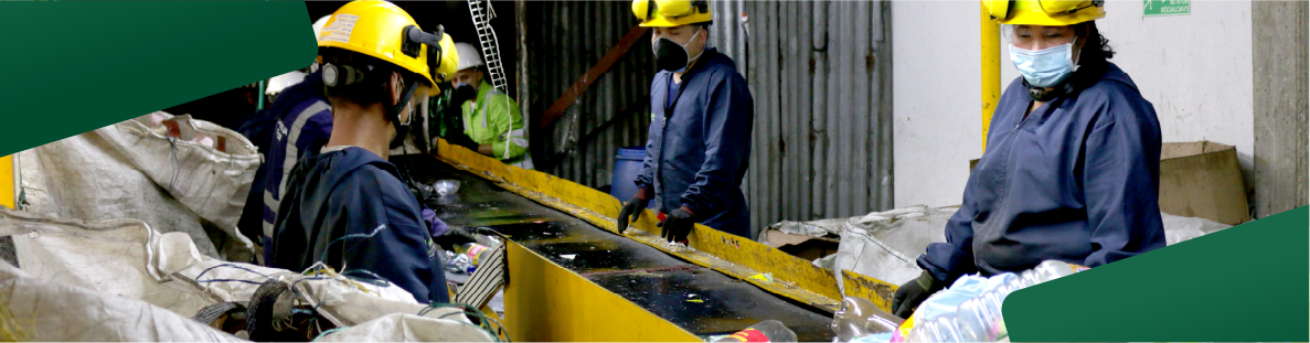 En la imagen: Tres recicladores de uniforme azul con cascos amarillos trabajan en una máquina, cinta sin fin, en la clasificación de plástico en una bodega de reciclaje.
