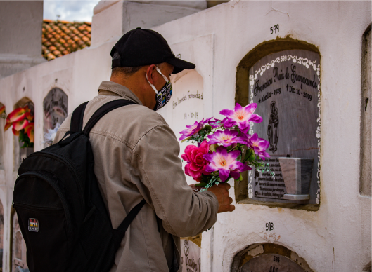 En la imagen: Un hombre con gorra negra, chaqueta y un morral en su espalda, vive un momento íntimo frente a un ser querido que falleció. Está de pie frente a una lápida color gris. En sus manos lleva un ramo de flores lilas, blancas y rojas. 