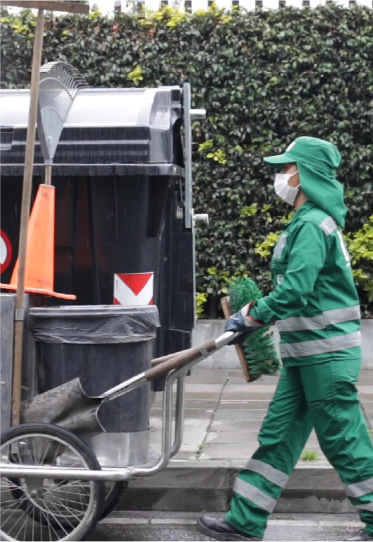 En la imagen: En primer plano, sobre la calle un operador con uniforme y con gorra de color verde, empuja un carrito que contiene bolsas, herramientas escobas y recogedor. De fondo un contenedor para depositar residuos. 