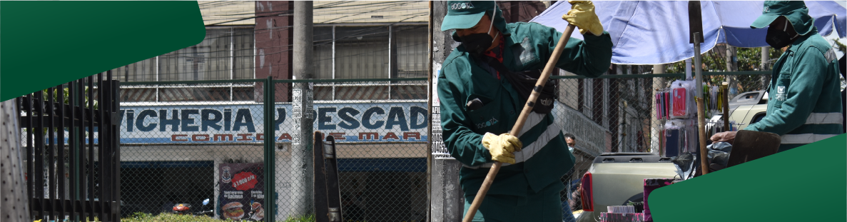 En la imagen: Dos operadores con uniforme verde oscuro y gorra con escoba en mano, barren la calle. De fondo locales comerciales.