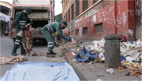 En la imagen: Dos operadores de recolección de residuos limpian en la calle una gran cantidad de residuos que fue esparcida sobre un andén, de fondo un camión recolector  está estacionado sobre la vía.
