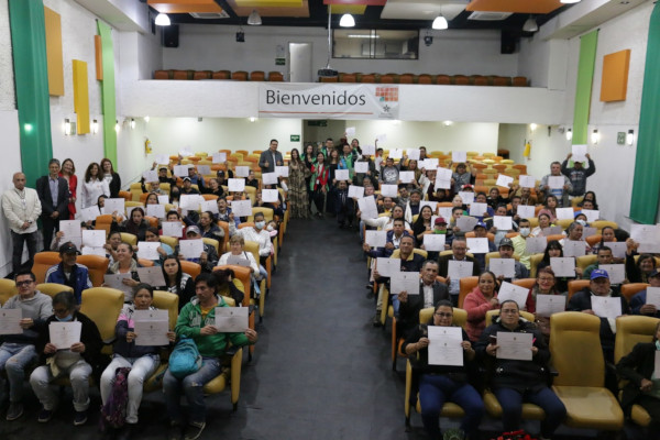  Foto al interior. Plano general de un auditorio en el que se ven personas sentadas a los dos lados levantando un diploma y al fondo se ven personas posando para la foto. Las paredes son blancas y cuenta con algunos detalles de colores verde, naraja, amarillo y verde.