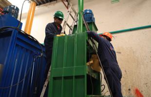 Dos hombres vestidos de azul, con casco naranja y verde, se encuentran sobre una escalera, manipulando una máquina.