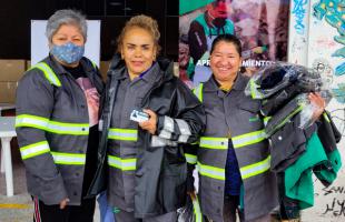 La foto es en una fachada (exterior) de fondo se ven a tres mujeres recicladoras que portan los nuevos uniformes y en sus manos tienen, más bolsas con uniformes y el carné RURO.