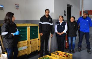 Dos profesionales que se encuentran al lado de una compostera (caja amarilla con verde) están dialogando con 3 personas. En frente tienen cananastas con residuos de frutas y verduras. 