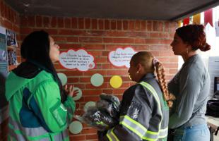 Una profesional de la UESP con la chaqueta verde habla con dos mujeres recicladoras entorno a unos afiches