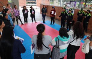 Mujeres escuchan atentamente a mujeres miembros de la policía. En el piso hay un tapete de color azul y rojo. En el fondo se ven paredes (adrillo).