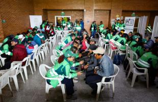 Foto al interior de un auditorio. Se ven seis filas de sillas ocupadas por recicladores de oficio quienes están siendo atendidos por profesionales de la UAESP que portan chaqueta verde