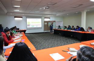 Grupo de personas reunidas en un auditorio. Las mesas están ubicadas en L, su mantel es de color naranja y las personas están observando la pantalla que proyecta la dinámica a desarrollar en la actividad. 