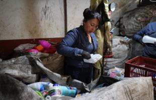 Foto en el interior de una bodega. En el fondo se ve una pared blanca con rojo, a su lado están bolsas con material reciclable. Se ven empaques plásticos y canastas en donde se ubican estos materiales.