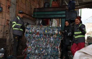  Foto en el interior de una bodega se encuentran dos mujeres y un hombre, recicladores de oficio, posando para la foto con una material plástico compactado y una máquina de compactar. . De fondo