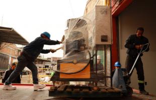 Es una fotografía de día. Y se puede ver dos hombres en plano general están sobre una grúa en la parte externa de una bodega, moviendo  maquinaria de procesamiento de plástico con un cargador de color azul.