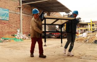 Es una fotografía de día. Y se puede ver a dos hombres con casco de protección de color azul, en plano general que están sobre una grúa en la parte externa de una bodega, trasladando una máquina.