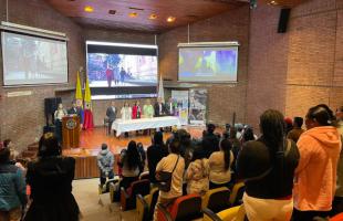 Foto al interior de un auditorio en contrapicada. Personas de pie en la mesa principal, el atril y sillas del auditorio. 