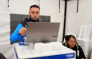 Foto al interior de una carpa blanca. En ella se ve a un joven de no más de 40 años, tiene puesta una chaqueta color azul cyan con negro y en sus manos tiene un computador portátil apoyado sobre una caja de impresora. Al lado derecho se ve a una mujer recicladora que está sentada y detrás de ella unas sillas de plastico, blancas.