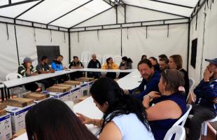 Foto al interior de una carpa blanca. Se ve mesas blancas formando una L. Alrededor están recicladores sentados haciendo parte de la reunión. En la parte central de la mesa, se ven unas cajas con computadores e impresoras.