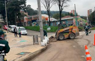 En una intersección vial, una máquina minicargadora y trabajadores realizan labores de limpieza y mantenimiento del espacio público. 