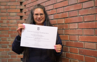 Una señora vestida de negro que usa gafas sostiene en sus manos un diploma. 