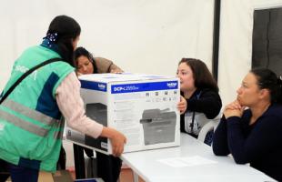 En el fondo de una carpa blanca están recicladoras alrededor de la mesa y hay se ve a una profesional de la UAESP haciéndole entrega de una caja con una impresora