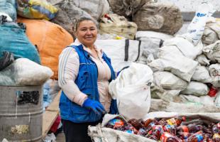 Foto de día en el interior de una bodega de reciclaje en la que se ven residuos almacenados en bolsas. En un plano 3/4 se ve a una mujer (representante legal de la Organización)  que está vestida con chaleco azul y sueter rosado y pantalón negro.