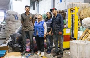 Foto de día en el interior de una bodega de reciclaje en la que se ven residuos almacenados en bolsas. Una máquina a uno de estos lados. En en el centro de la foto hay dos mujeres y dos hombres