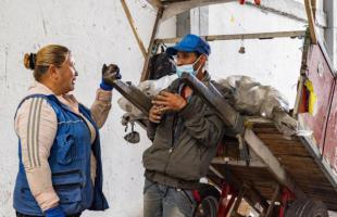 Foto de día en el interior de una bodega de reciclaje en la que se ven residuos almacenados en bolsas. Una carreta de fondo y un carretero interactuando con una señora.