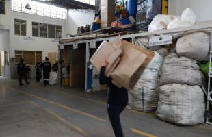 Es de día - Varias mujeres están en una Bodega de reciclaje. Se ven lonas color blanco con productos dentro. Una mujer con uniforme azul oscuro va llevando cartones sobre su hombro.