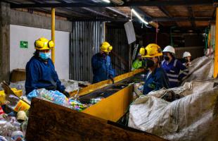 Foto en exterior en el que se ven a 6 recicladores trabajando en la selección de material y tienen cerca a una banda transportadora de materiales. Los recicladores portan uniforme azul oscuro y casco con tapabocas como equipos de protección