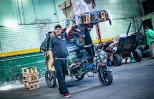 Foto en el interior de un CTCC Los Mártires. Reciclador vestido de color azul oscuro con una mano levantada sujetando su bicicarreta. En el fondo se ven residuos arrumados al fondo. Y una pared verde oscuro con verde claro