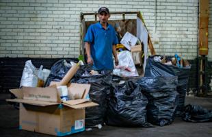 Foto en el interior de un CTCC Los Mártires. Reciclador con camisa polo azul claro, jean y gorra. En el fondo se ve una pared blanca y justo detrás de él se ve una carreta con residuos. En la parte delantera se ven bolsas negras y algunas cajas con residuos