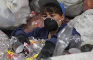 Foto al interior de una bodega. Plano de la foto en detalle. Se ve una señora con overol y gorra azul. Cuenta con protección (guantes, gorra y tapabocas). La recicladora está manipulando y separando residuos de plástico.