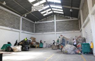 Foto de día, del interior de centro transitorio de Usaquén. Es una foto de día. Las paredes son con ladrllo y color blanco. El techo contempla tejas traslucidas. En el interior se encuentran 4 carreteros junto a sus carretas haciendo separación de material.