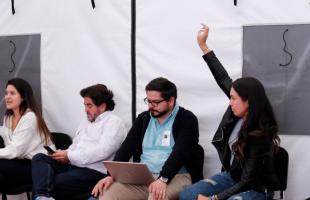 Cuatro personas sentadas participando de la Mesa de Plástico. Una de ellas, de chaqueta negra está levantando la mano pidiendo la palabra. Las otras; una está mirando el computador, otra su celular y otra está interviniendo. 