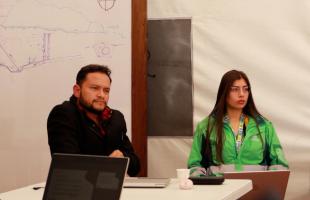 En un entorno cerrado de una carpa, están dos personas, una de ellas, una mujer está identificada con chaqueta de la UAESP. La otra persona es un hombre y tiene chaqueta negra. Los dos prestan atención a los participantes de la Mesa Industrial del Plástico.