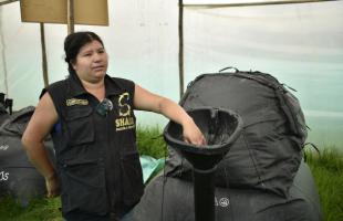 Plano general. Es de día. Una membrana negra está de fondo en el que una señora - recicladora de oficio, está tocando la campaña de la membrana. Alimentandola con residuos orgánicos. En el fondo se ve pasto verde y una polisombra.